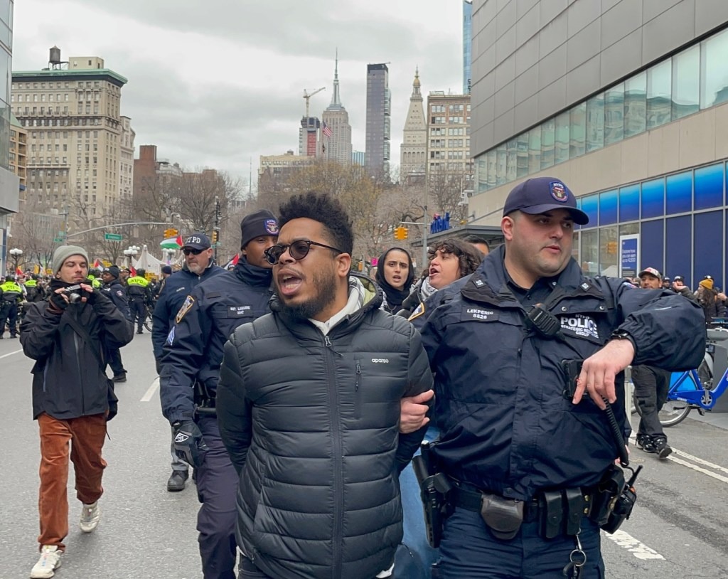 Arrest photo — Manolo De Los Santos in green sash restrained by NYPD in riot gear